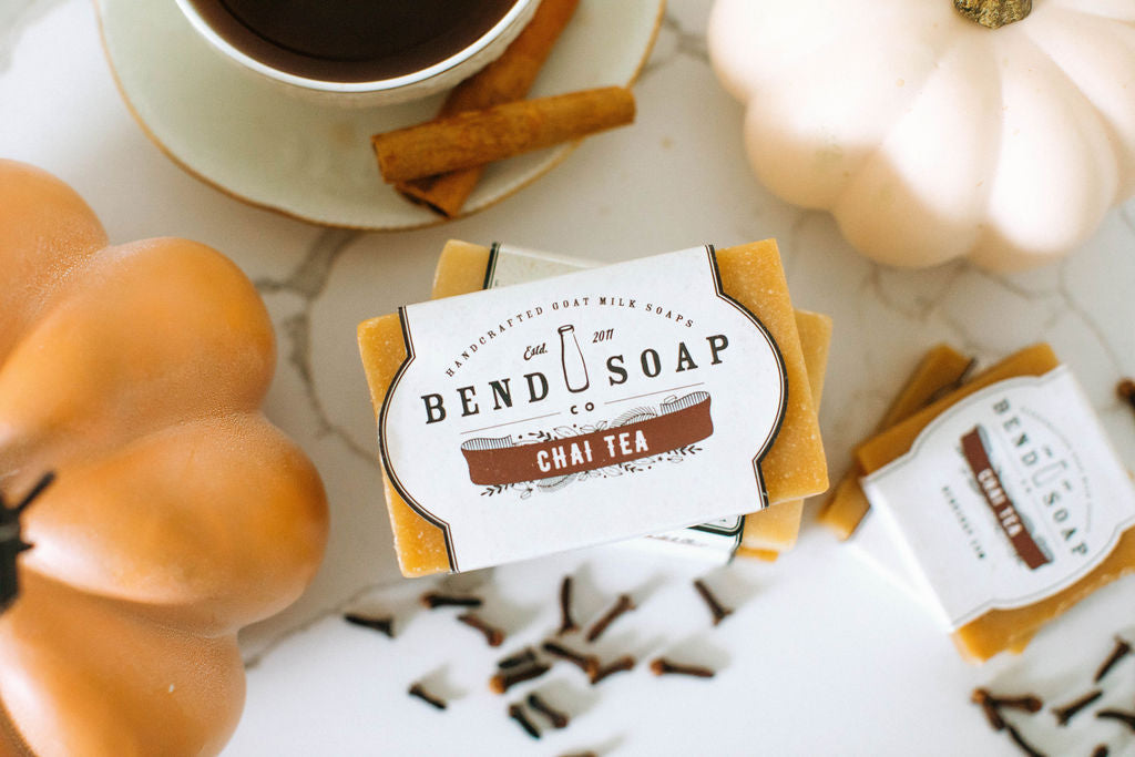Two orange pumpkins, a cup of tea, cinnamon sticks, and Chai Tea Goat Milk Soap bars labeled “Chai Tea Goat Milk Soap” rest on a white surface with scattered cloves, creating a cozy autumn scene filled with warm cinnamon chai aroma.