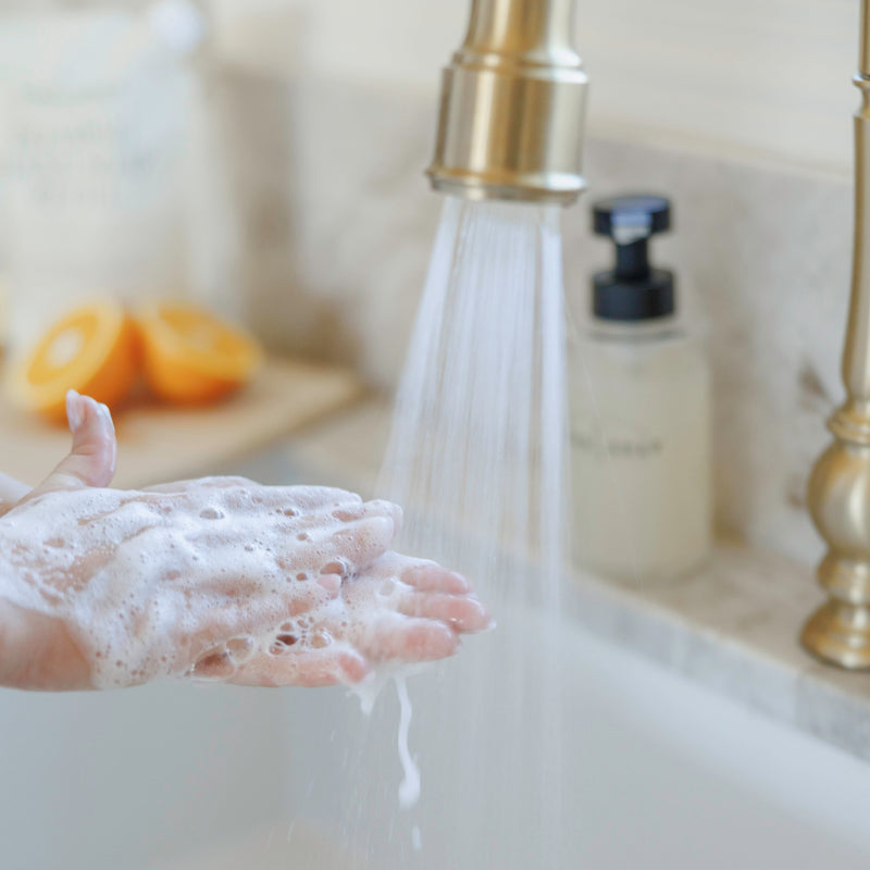 Hand under a running faucet with soap suds, next to a soap dispenser on a bathroom counter.