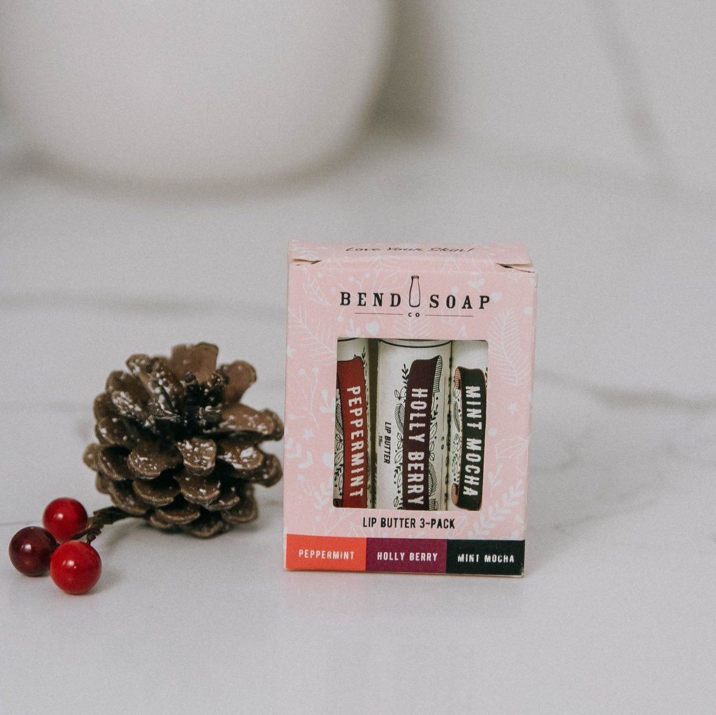 A box of Lip Butter Holiday Trio—featuring peppermint, holly berry, and mint mocha—is placed by a pine cone with red berries on a light surface.