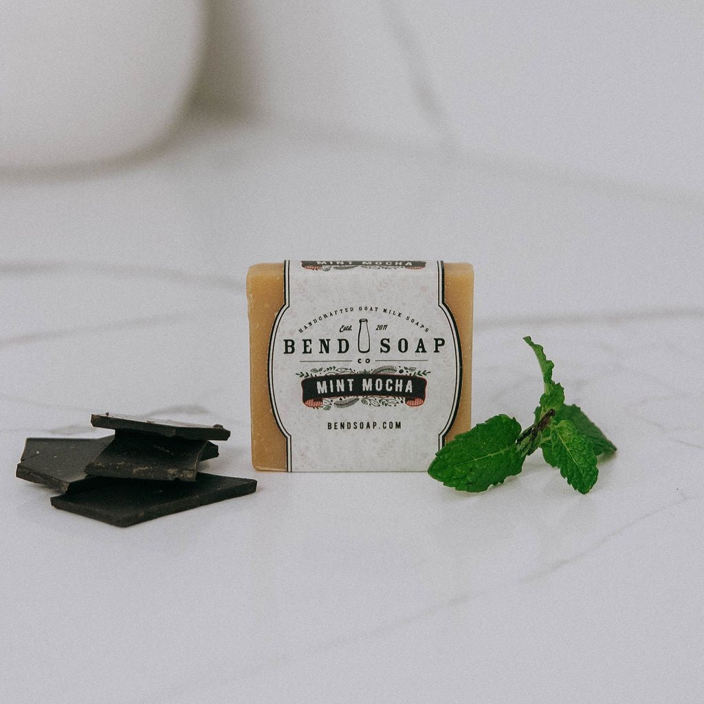 A Mint Mocha Goat Milk Soap bar rests on a white surface, flanked by dark chocolate pieces on the left and fresh mint sprig on the right—a limited edition holiday classic.
