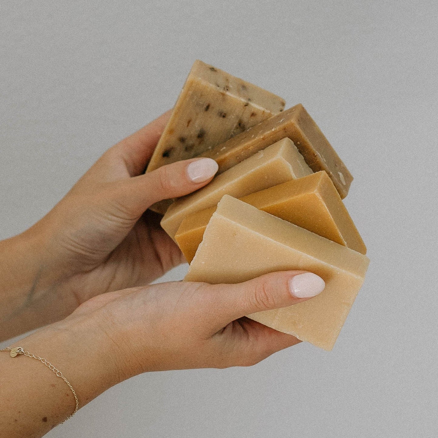 A person with light pink nail polish holds several natural, handmade soap bars in different earthy tones against a neutral background.