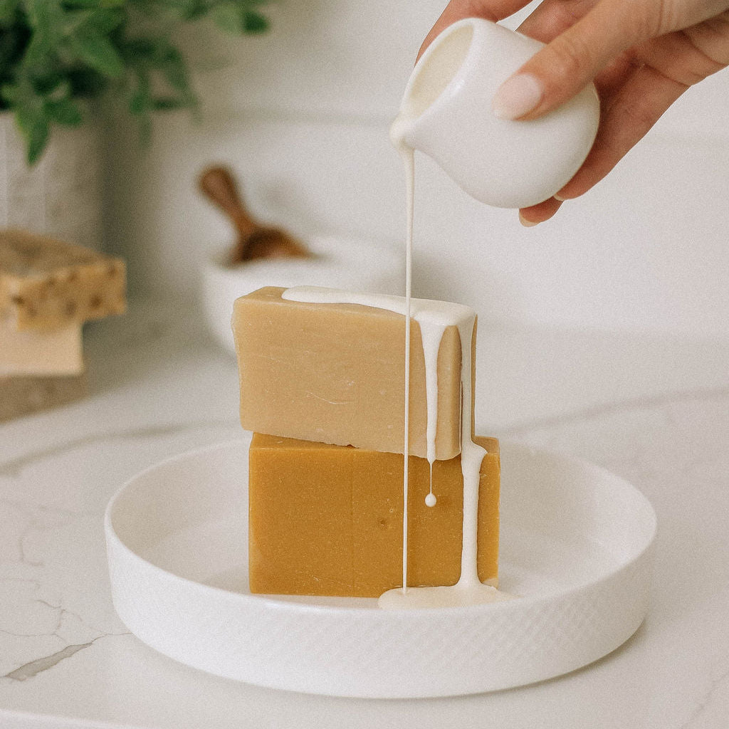 A hand pours a creamy white liquid over two stacked soap bars—one tan, one golden—on a white dish, elegantly showcasing Vienna Rose Goat Milk Soap Scrap (1 lb.) with a soft backdrop of plants and a soap dish.