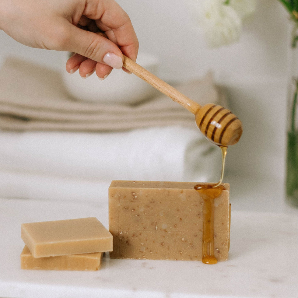 A hand drizzles honey over an Oatmeal and Honey Goat Milk Soap Scrap (1 lb.), perfect for sensitive skin, with another soap and folded towels in the background.