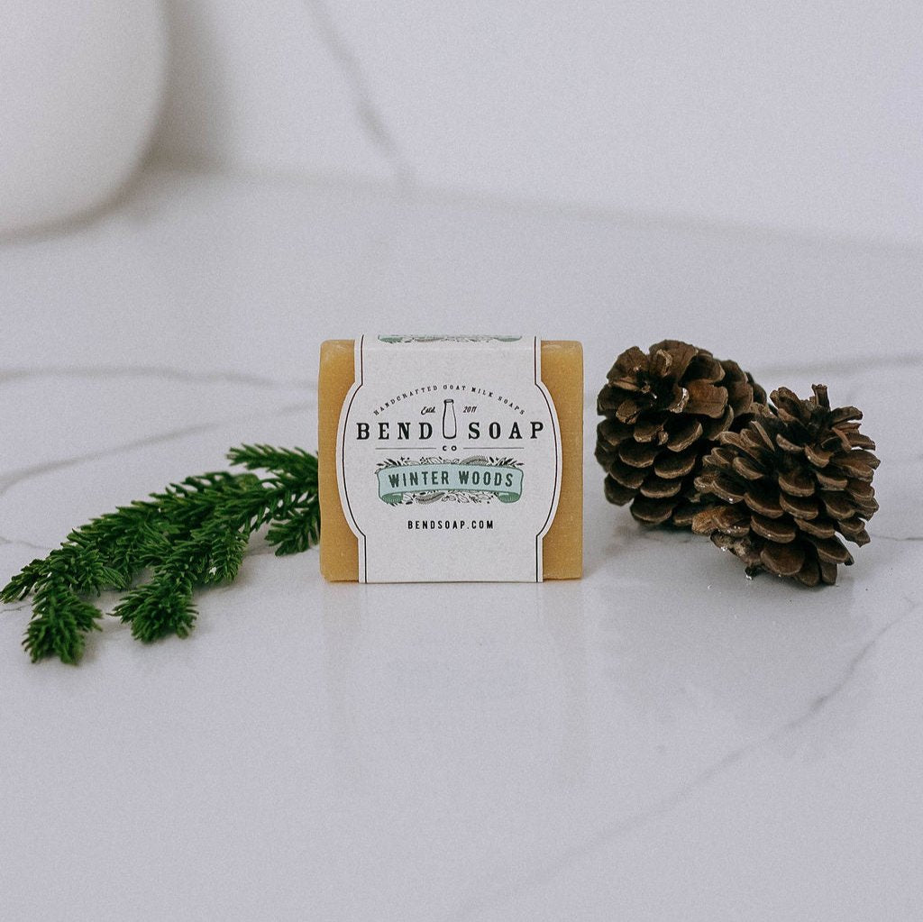 The Winter Woods Goat Milk Soap bar rests on a white surface beside two pinecones and a green pine branch, capturing the season's essence with pure essential oils.