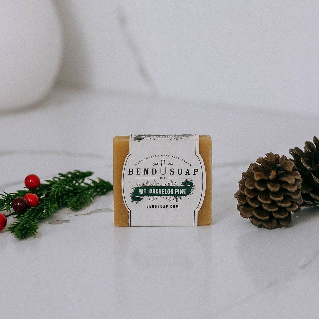 A bar of Bend Soap labeled "Mt. Bachelor Pine" is placed on a white marble surface, with a pine cone and a sprig of pine with red berries beside it.