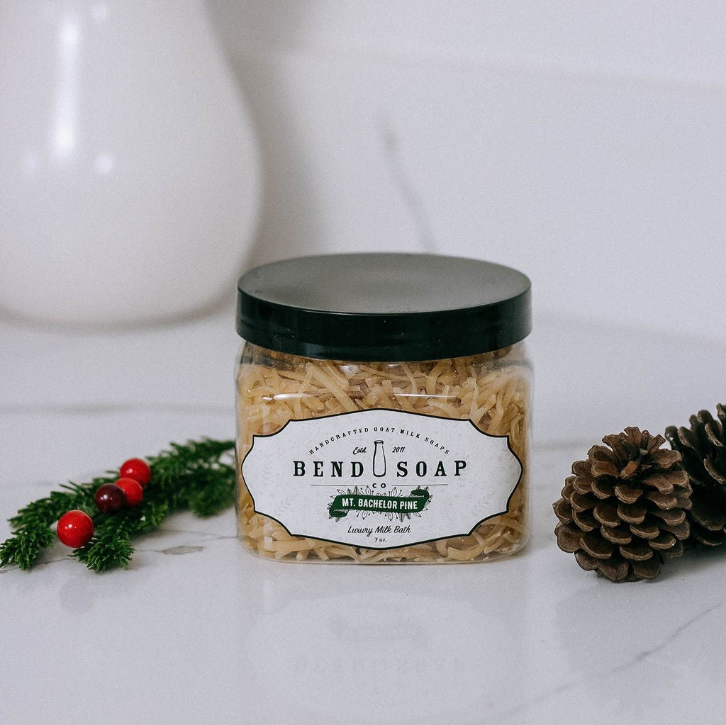 A clear jar of Bend Soap Co. Mt. Bachelor Pine luxury milk bath sits on a white surface next to a pine cone and a small sprig of pine with red berries.