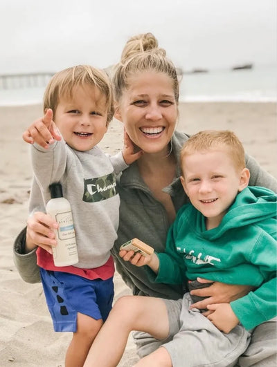 a young mother and her two sons on a beach holding a bottle of goat milk lotion